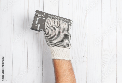 Tool. Hand in glove is holding industrial stapler on a white wooden background .