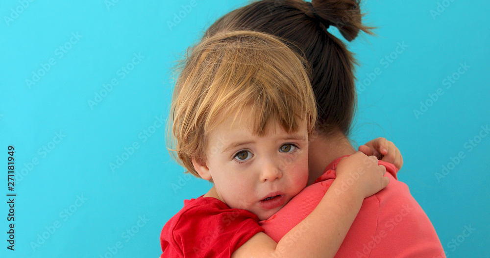 Side view of young mom cuddling sad kid looking at camera isolated on blue background Side view of young mom cuddling sad kid looking at camera isolated on blue background
