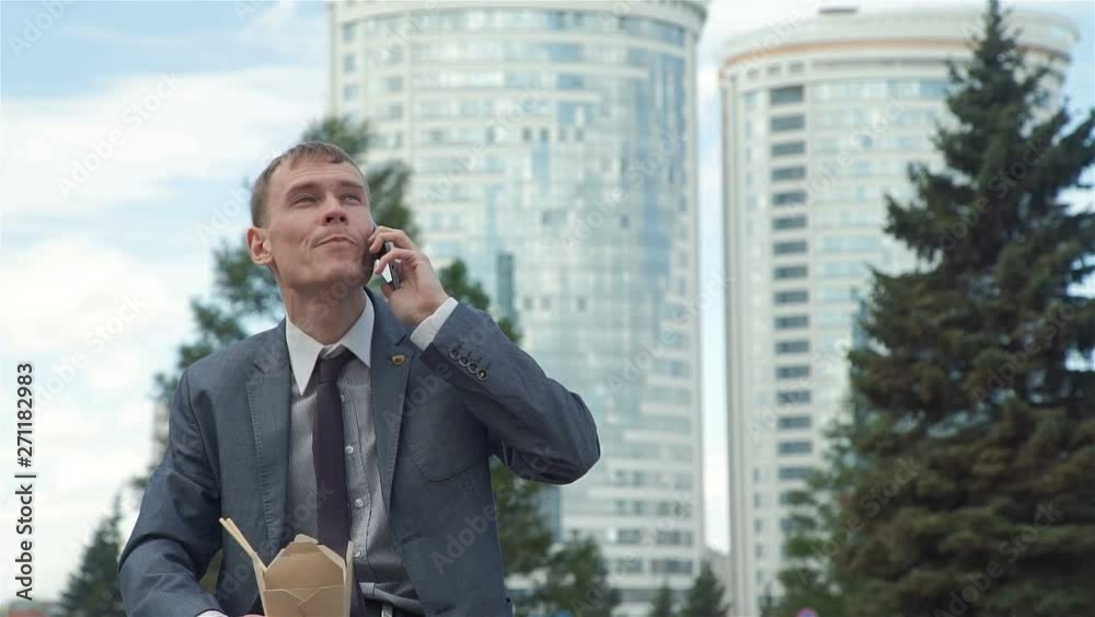 Handsome Businessman Sitting on a Bench and Talking on Phone while Having Lunch Outdoors in Slow Motion. He is Holding a Cardboard Box with Chinese Noodles in his Hands. Lifestyle and Business Concept Handsome Businessman Sitting on a Bench and Talking on Phone while Having Lunch Outdoors in Slow Motion. He is Holding a Cardboard Box with Chinese Noodles in his Hands. Lifestyle and Business Concept