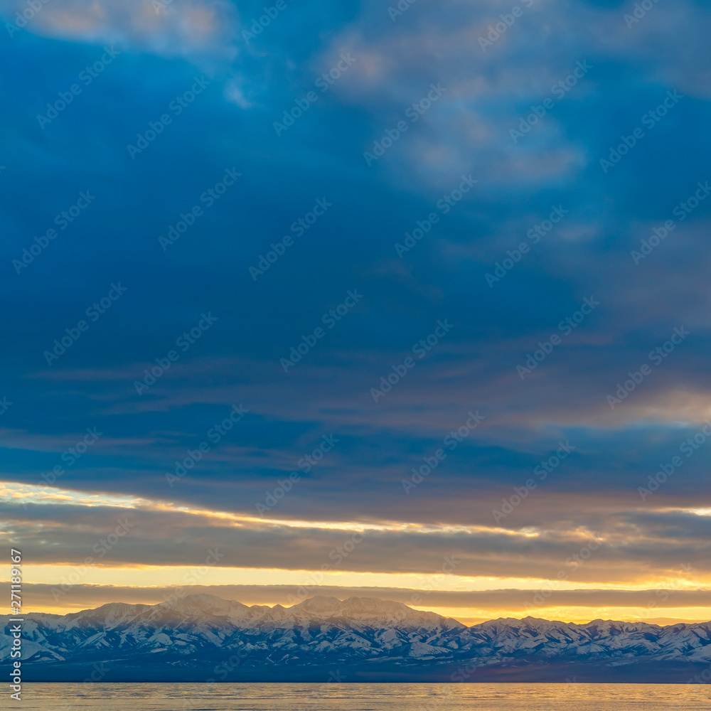 Fototapeta premium Square frame Scenic panorama of a lake and snow capped mountain under a striking cloudy sky