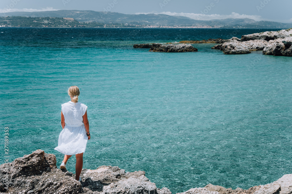 Jung adult women in white dress on summer vacation in front of sea coast landscape of small beach with crystal clear blue water. Greece, Kefalonia