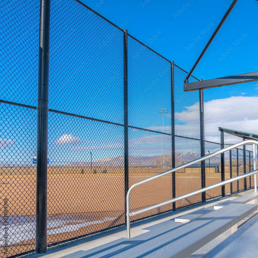 Square Sunlit bleachers overlooking a vast sports field on the other side of the fence Square Sunlit bleachers overlooking a vast sports field on the other side of the fence