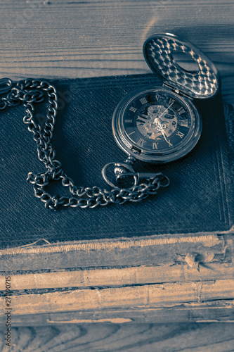 Antique pocket fob watch with a chain on a stack of old books. On a wooden background with colour toning.