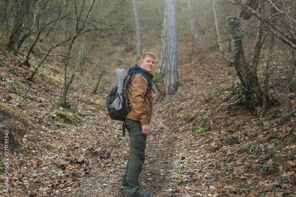 Redhead man in a brown-and-blue windbreaker with backpack stands sideways and looks at the camera in a forest in the Crimea in the early spring. Travel, adventure, hiking and motivation concept.