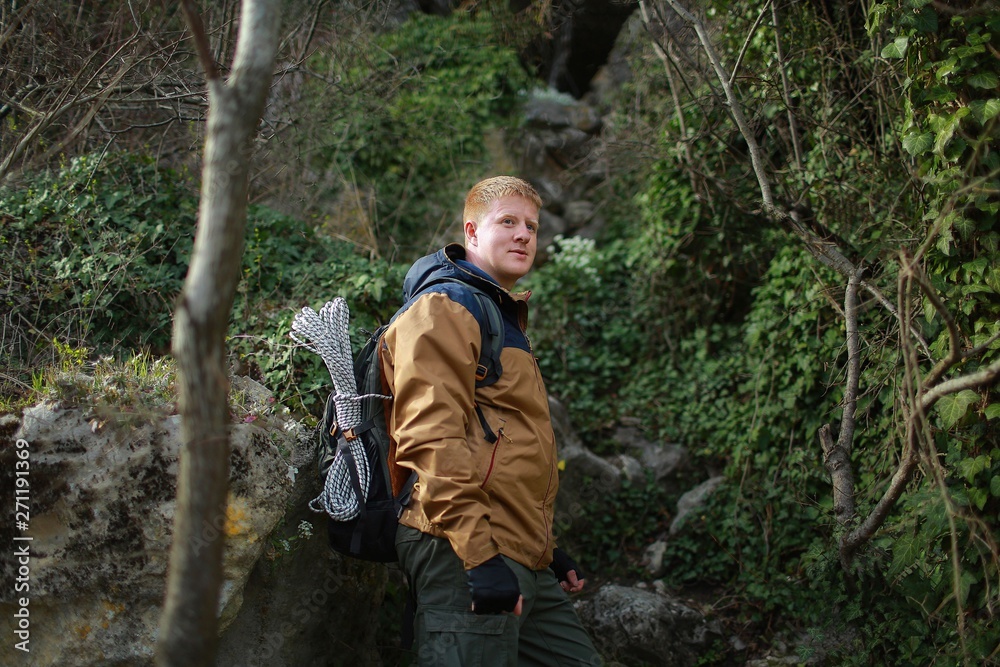 Naklejka premium Redhead man in a brown-and-blue windbreaker with backpack and rope stands in mountain forest in the Crimea in the early spring. Travel, adventure, hiking and motivation concept.