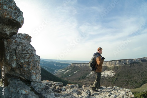 Redhead man in a brown-and-blue windbreaker stands in cave town Mangup-Kale on the edge of a cliff on the mountain plateau in the Crimea. Travel, adventure, climbing and hiking concept.