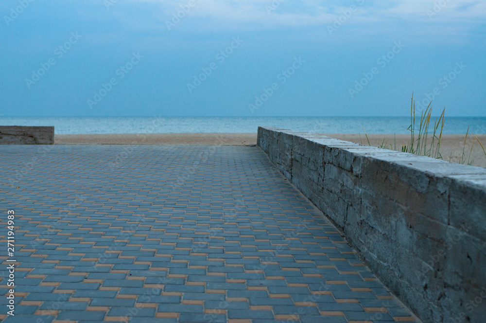 Chess pattern walkway near beach. Tranquil seascape in dusk with nobody and cement border between walkway and beach sand with high wild plants. Blurred moon in blue evening sky. Black sea landscape 