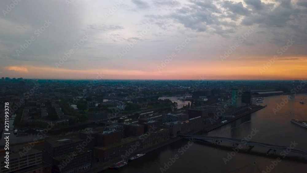 Flying over Amsterdam skyline by sunset by drone over the IJ river near central station with beautiful red sky and white train heading to train station. With freight boats in the reflecting waters Flying over Amsterdam skyline by sunset by drone over the IJ river near central station with beautiful red sky and white train heading to train station. With freight boats in the reflecting waters