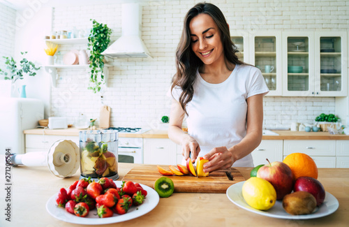 Beautiful woman making fruits smoothies with blender. Healthy eating lifestyle concept portrait of beautiful young woman preparing drink with bananas, strawberry and kiwi at home in kitchen.