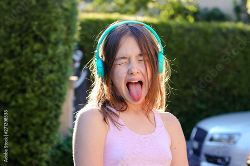 Close-up portrait of funny preteen girl with wireless headphones on head with closed eyes and open mouth with stuck out tounge. Teenager age emotions and protest