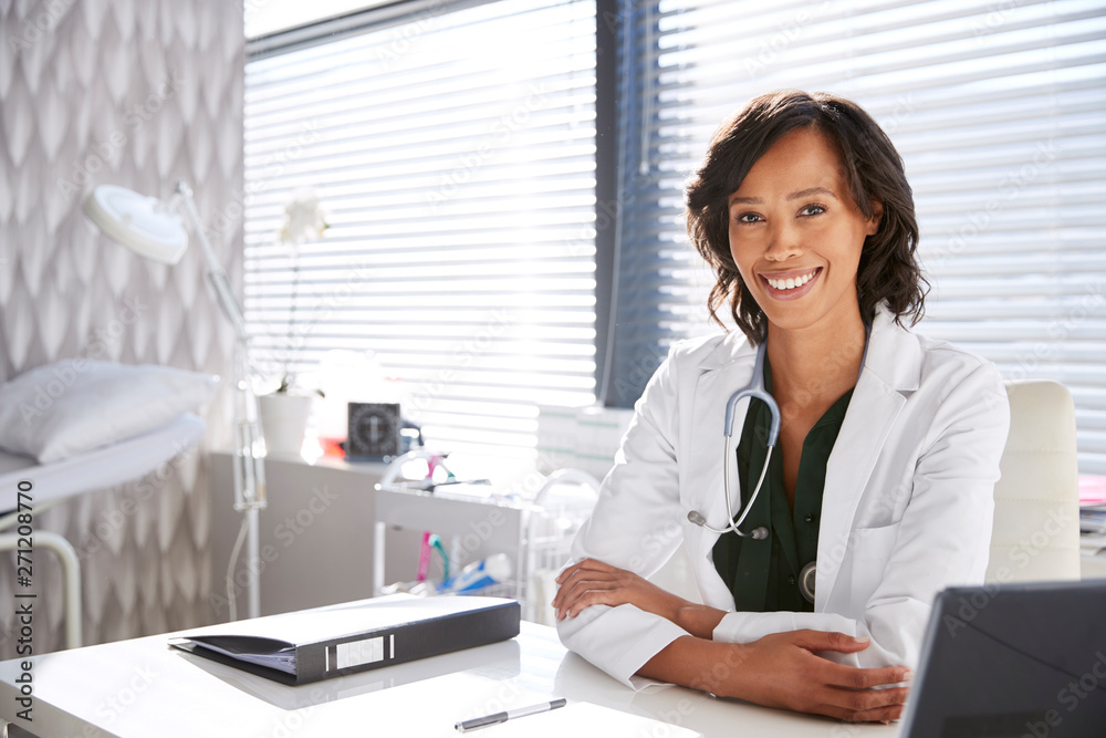 Portrait Of Smiling Female Doctor Wearing White Coat With Stethoscope Sitting Behind Desk In Office Portrait Of Smiling Female Doctor Wearing White Coat With Stethoscope Sitting Behind Desk In Office