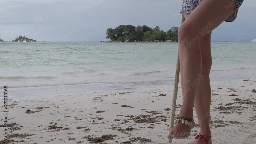 Blonde woman swinging legs on beach swing Anse Volbert Cote d'Or, Praslin, Seychelles, Indian Ocean. Islet of Chauve Souris on background. Lifestyle female funny, summer vacation. SLOW MOTION