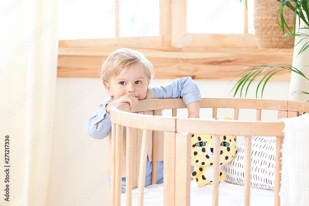 A boy stands alone beside a cot in the nursery. Lonely baby is in kindergarten near the crib. Loneliness. Eco-friendly children's room decor in the Scandinavian style. The boy is at home. interior A boy stands alone beside a cot in the nursery. Lonely baby is in kindergarten near the crib. Loneliness. Eco-friendly children's room decor in the Scandinavian style. The boy is at home. interior