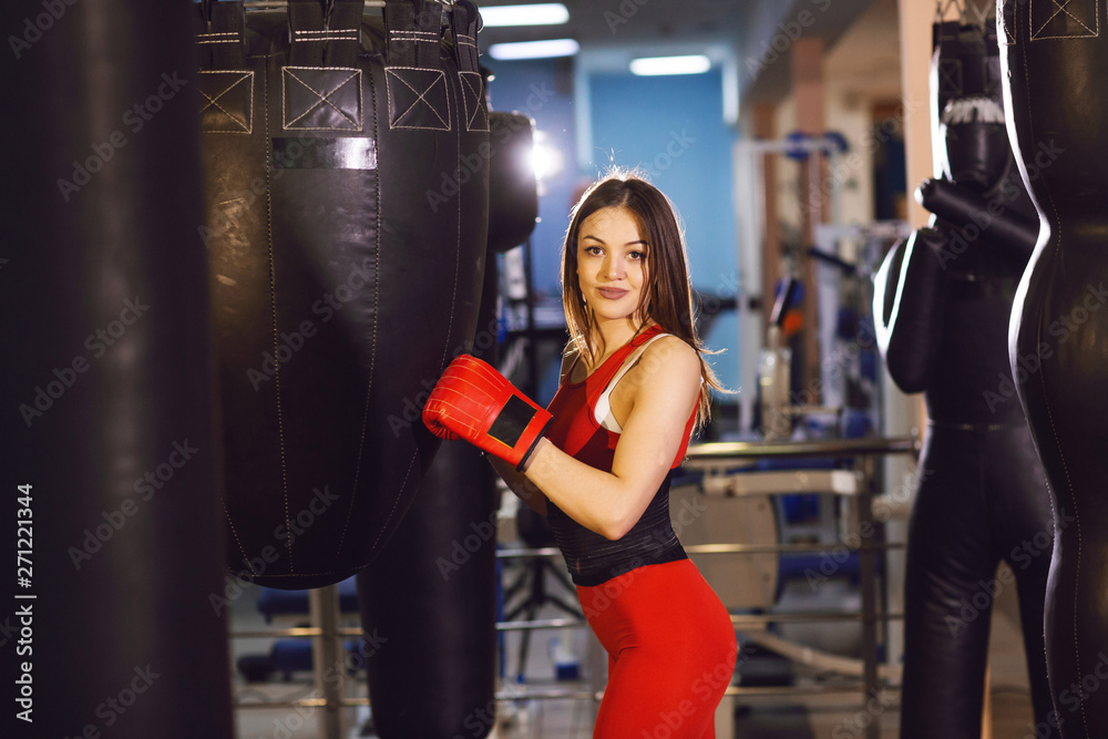 Young woman in red sports clothes and boxing gloves, trains with a boxing pear in a dark gym