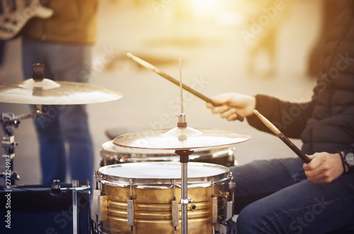 A street musician, dressed in a black jacket and blue jeans, with a watch on his hand, plays a drum set with old battered drumsticks in the sunlit street. photo