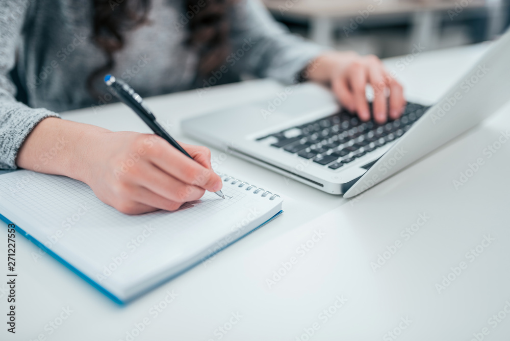 Close up of woman's hands writing in spiral notepad and using laptop. Close up of woman's hands writing in spiral notepad and using laptop.
