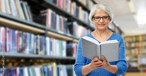 vision, wisdom and old people concept - smiling senior woman in glasses reading book over library background