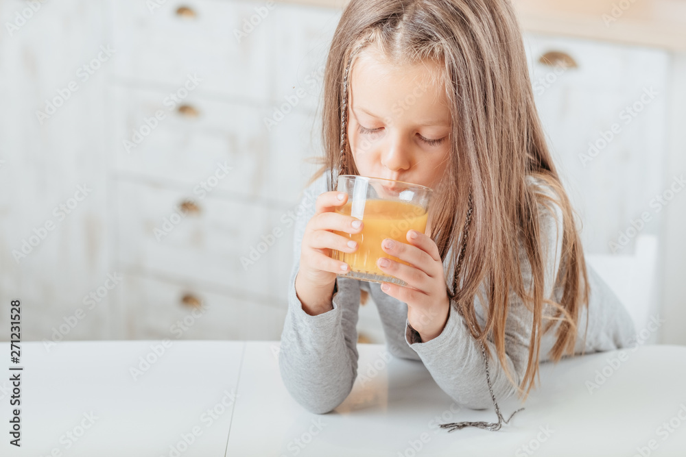 Healthy morning beverage. Cute little girl drinking orange fruit juice in kitchen. Blur background. Copy space. Healthy morning beverage. Cute little girl drinking orange fruit juice in kitchen. Blur background. Copy space.