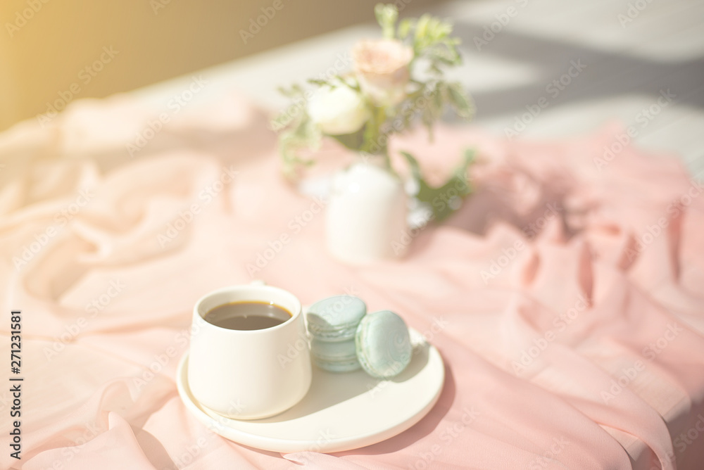 French macaroon blue plate on the pink and pink coffee cup standing on a wooden table with a pink tablecloth white vase with flowers roses and greens French macaroon blue plate on the pink and pink coffee cup standing on a wooden table with a pink tablecloth white vase with flowers roses and greens