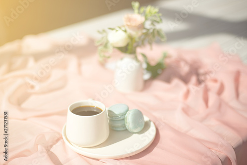 French macaroon blue plate on the pink and pink coffee cup standing on a wooden table with a pink tablecloth white vase with flowers roses and greens