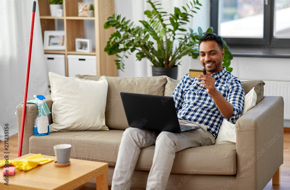household, housework and technology concept - indian man with laptop computer and credit card shopping online after home cleaning household, housework and technology concept - indian man with laptop computer and credit card shopping online after home cleaning