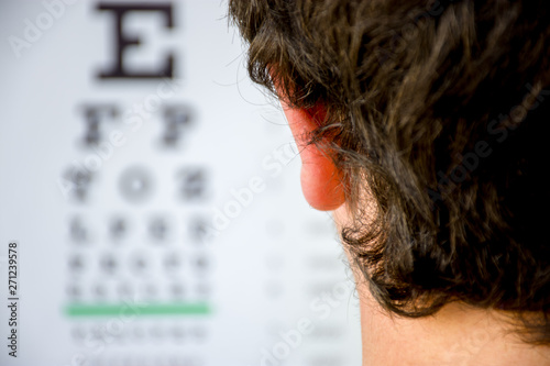 Concept photo of myopia or nearsightedness as diseases of eye and the optical system. In the background blurry fuzzy table for testing visual acuity, in the front - head of the person in focus closeup