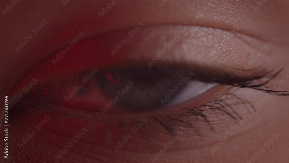 Close-up shoot of light-blue eye blinking with reflection of lamp on it in red light.