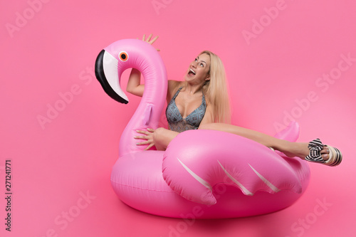 Picture of blonde in bathing suit and in pink glasses sitting on inflatable flamingo on empty pink background
