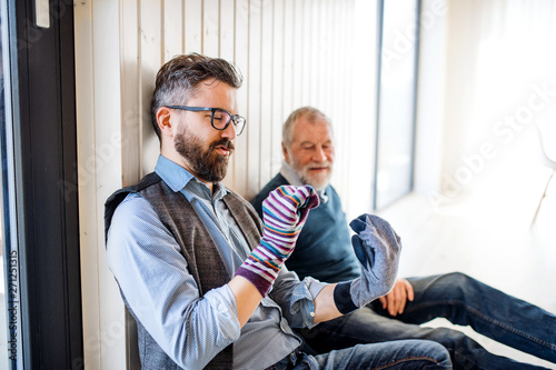 Portrait of adult hipster son and senior father sitting on floor indoors at home, having fun.