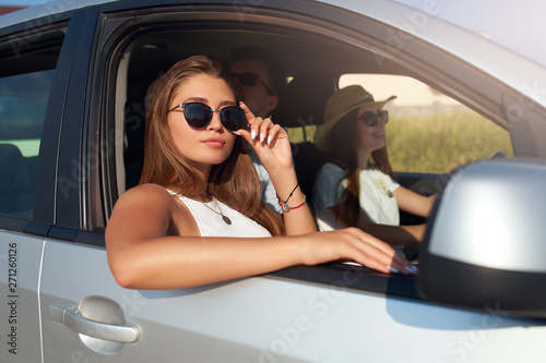Group of friends rented a car on summer road trip and arrived to the sea beach. Woman in glasses looks out of the car window. Passanger girl having fun with friends in vehicle. Travel lifestyle. © artiemedvedev