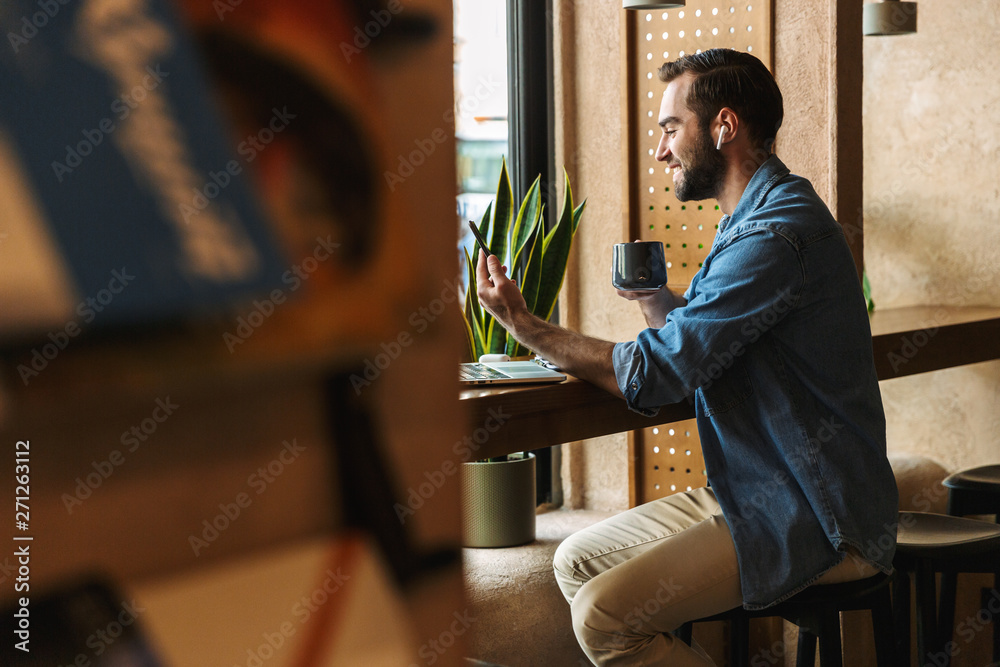 Photo of unshaven caucasian man using earpod and cellphone with cup of tea while working in cafe indoors