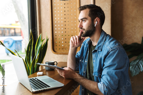 Fényképezés Photo of brooding caucasian man using earpod and clipboard with laptop while wor
