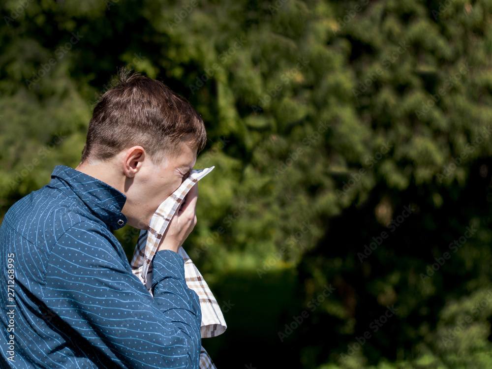 Closeup portrait of a young man with allergies. A man sneezes into a scarf on the background of green trees. Closeup portrait of a young man with allergies. A man sneezes into a scarf on the background of green trees.