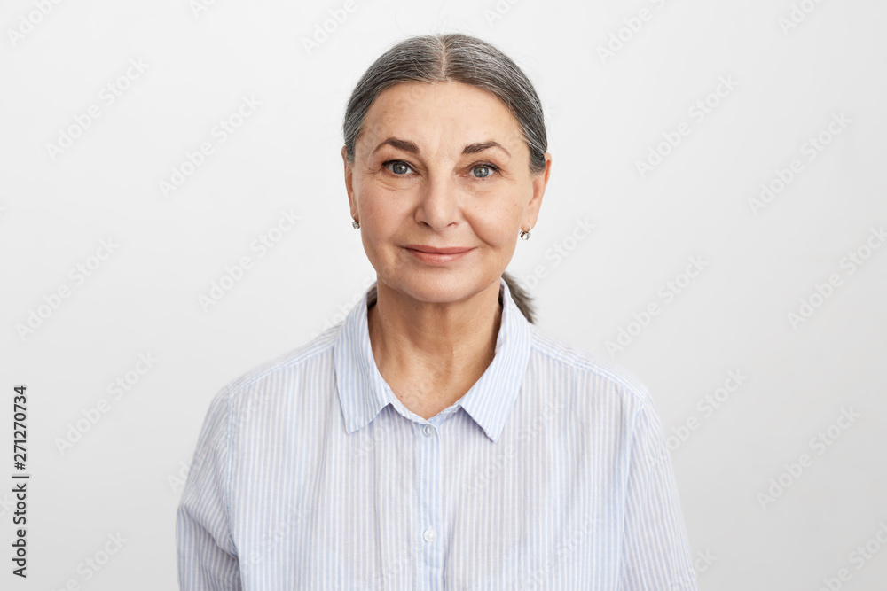Isolated image of attractive friendly looking mature senior female employee with wrinkles, gray hair and blue eyes wearing formal shirt smiling confidently at camera, being ready for working day Isolated image of attractive friendly looking mature senior female employee with wrinkles, gray hair and blue eyes wearing formal shirt smiling confidently at camera, being ready for working day