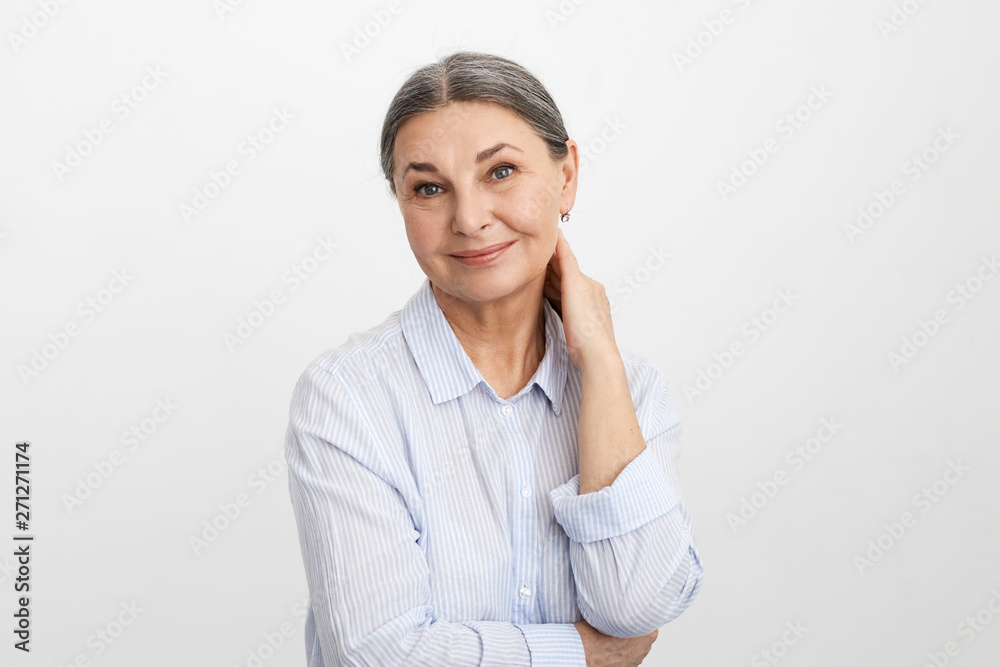 Gorgeous beautiful elderly female on retirement dressed in stylish blue shirt rubbing back of her neck and smiling happily at camera, posing agaisnt white wall background with copyspace for your text Gorgeous beautiful elderly female on retirement dressed in stylish blue shirt rubbing back of her neck and smiling happily at camera, posing agaisnt white wall background with copyspace for your text