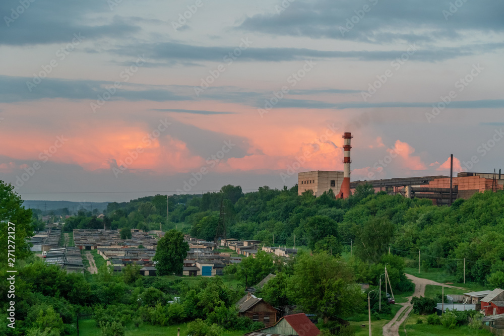 factory pipe with smoke against a background of pink sunset and green trees, garages on the outskirts of the city
