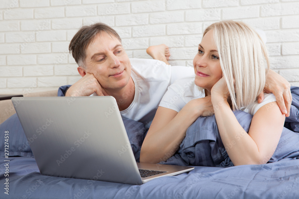 Smiling attractive couple lying on bed using laptop communicating online at home, happy man and woman typing on computer, enjoying internet shopping or chatting in social networks in bedroom.