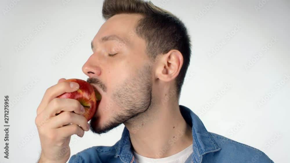 Portrait of young man with beard standing isolated on white background and eating red apple while looking at camera Portrait of young man with beard standing isolated on white background and eating red apple while looking at camera