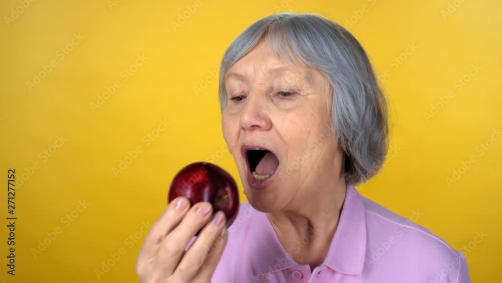Portrait of elderly woman with grey hair standing isolated on yellow background and eating delicious red apple while looking at camera