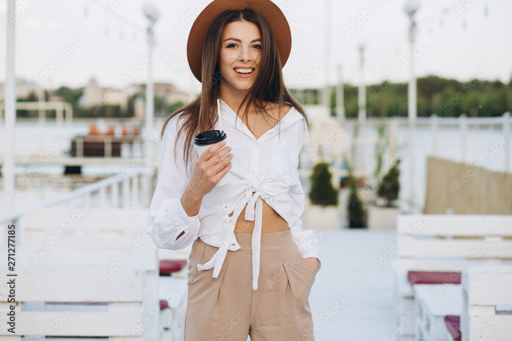 Fototapeta premium A stylish woman drinking coffee and walking along the beachfront in a warm summer day on the sunset