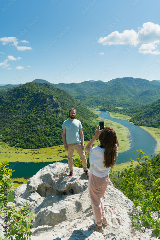 girl makes a photo of her boyfriend on the background of a magnificent landscape with a river and mountains