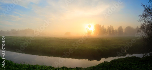 Serene summer panoramic landscape with forest river and meadow covered by fog at sunrise.