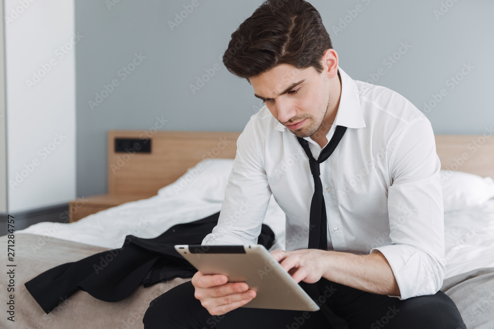Photo of handsome thinking businessman using tablet computer while sitting on bed in hotel apartment Photo of handsome thinking businessman using tablet computer while sitting on bed in hotel apartment