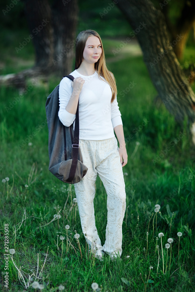 young woman smiling in white clothes came to nature to the lake to do yoga with a rug in a bag behind her back, among the trees at sunset young woman smiling in white clothes came to nature to the lake to do yoga with a rug in a bag behind her back, among the trees at sunset