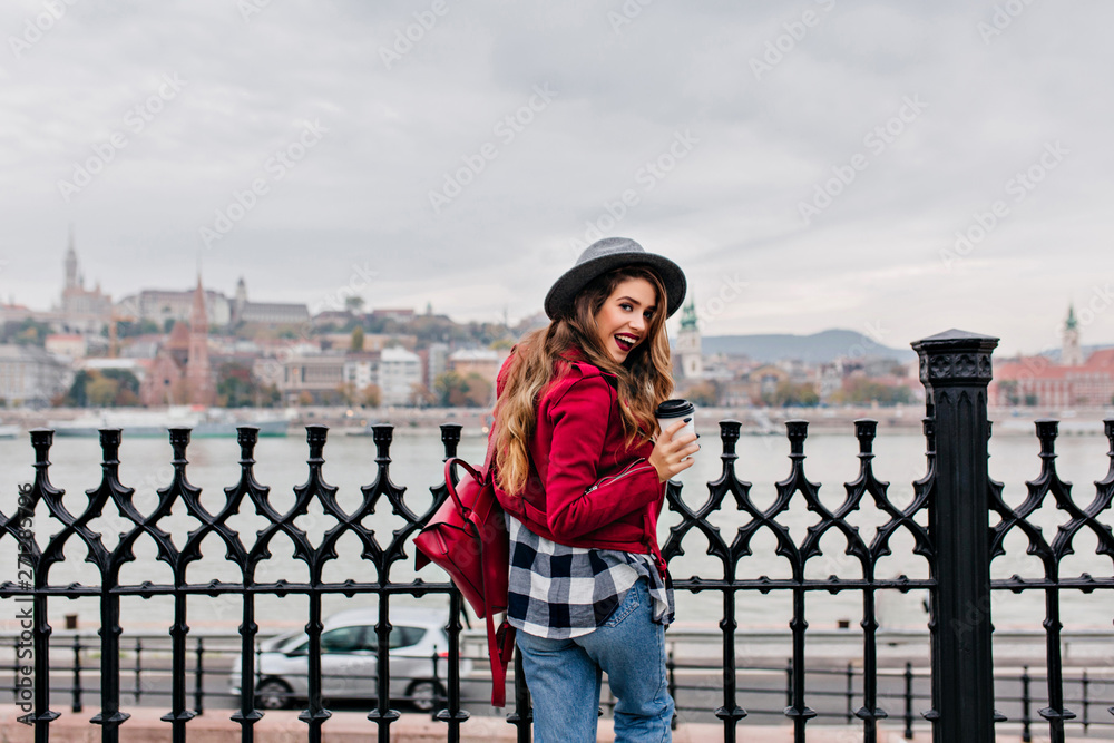 Photo from back of excited brunette lady in checkered shirt enjoying river views. Pleasant european girl in hat looking over shoulder and drinking coffee.