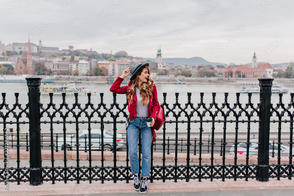 Full-length photo of good-looking girl with wavy hair posing at bridge. Slim stylish lady in vintage denim pants standing on city background.