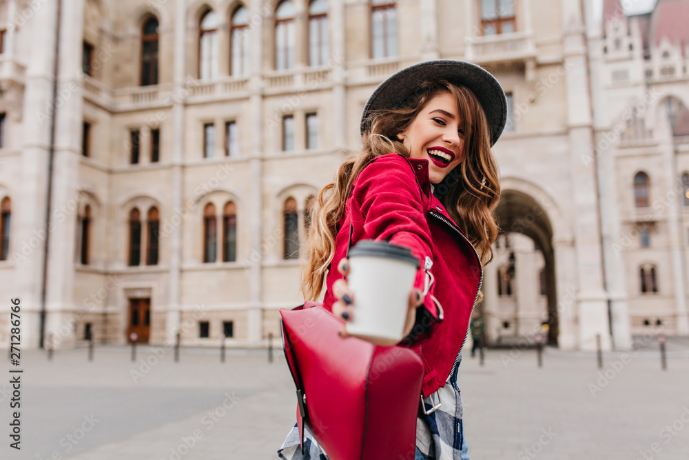 Outdoor photo of fashionable white woman posing with cup of latte on architecture background. Pretty long-haired brunette model expressing energy while walking around town.