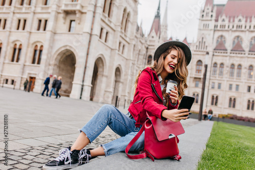 Attractive curly girl holding cup of coffee and making selfie on square in european city. Charming female traveler with backpack enjoying weekend in Europe and laughing.