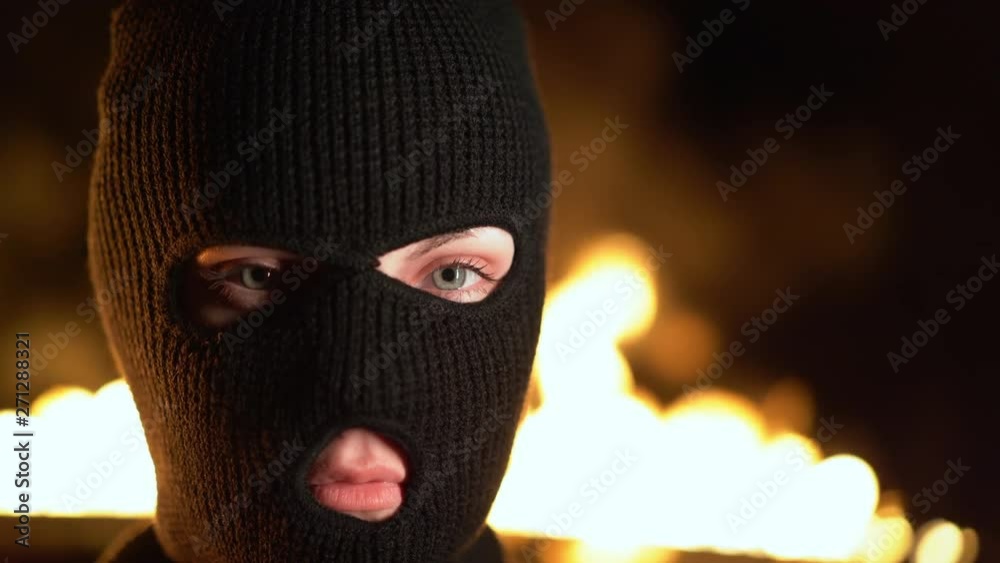 Portrait of young woman in black balaclava against backdrop of a blazing night fire. Concept of mass rallies and riots Portrait of young woman in black balaclava against backdrop of a blazing night fire. Concept of mass rallies and riots