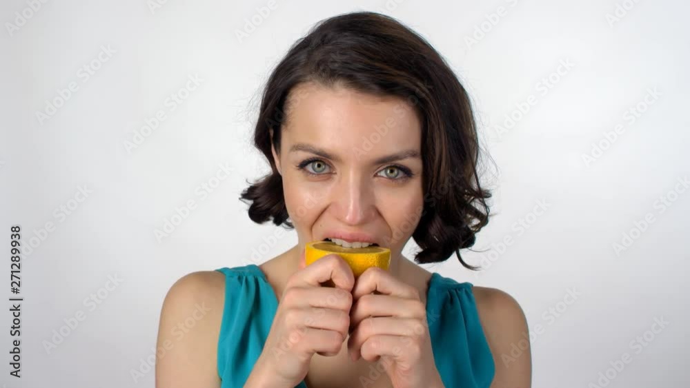 Portrait of woman standing isolated on grey background and biting juicy orange while looking at camera